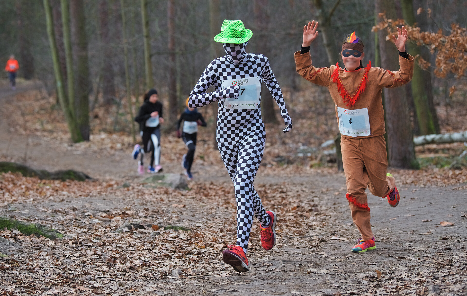 Berliner Silvesterlauf: Läufer:innen im Indianerkostüm sowie karierten Ganzkörper-Trikot mit Gesichtshaube auf der Strecke durch den Wald @ SCC EVENTS / Tilo Wiedensohler