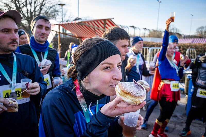 Eine Frau beißt glücklich in einen Pfannkuchen; Im Hintergrund weitere Teilnehmende des Laufs.