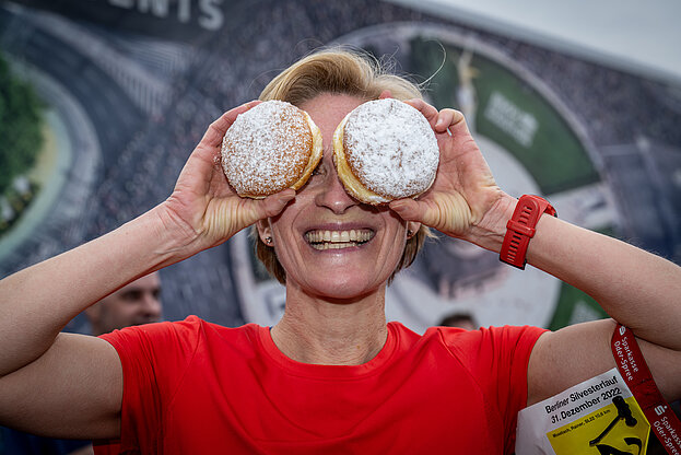 Berliner Silvesterlauf: Lachende frau in rotem T-Shirt hält sich zwei Pfannkuchen vor die Augen @ SCC EVENTS / Tilo Wiedensohler