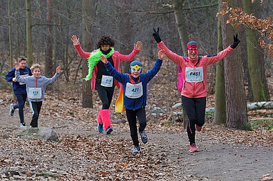 Spielbank Berlin Silvesterlauf 2019: Costumed family runs joyfully along a forest path with arms raised @ SCC EVENTS / Tilo Wiedensohler